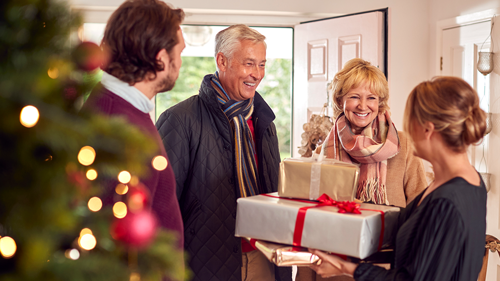 An elderly couple welcomes their family, while exchanging gifts near a Christmas tree.