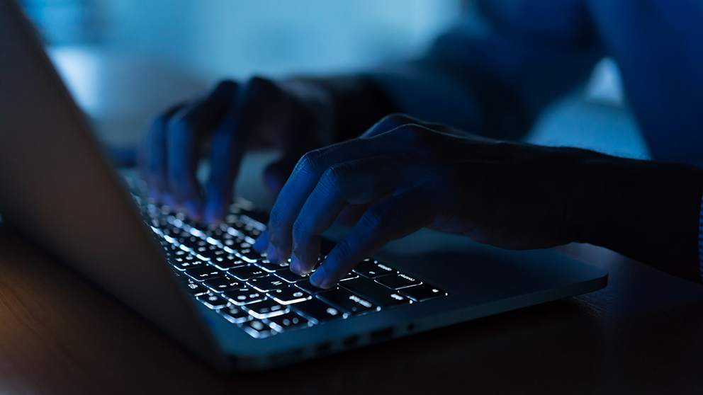 A pair of hands are illuminated by the light of a laptop keyboard in a dark room.