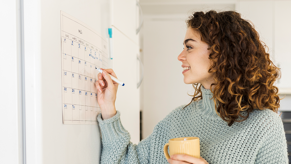 A woman with curly hair smiles as she writes on a calendar while holding a mug.