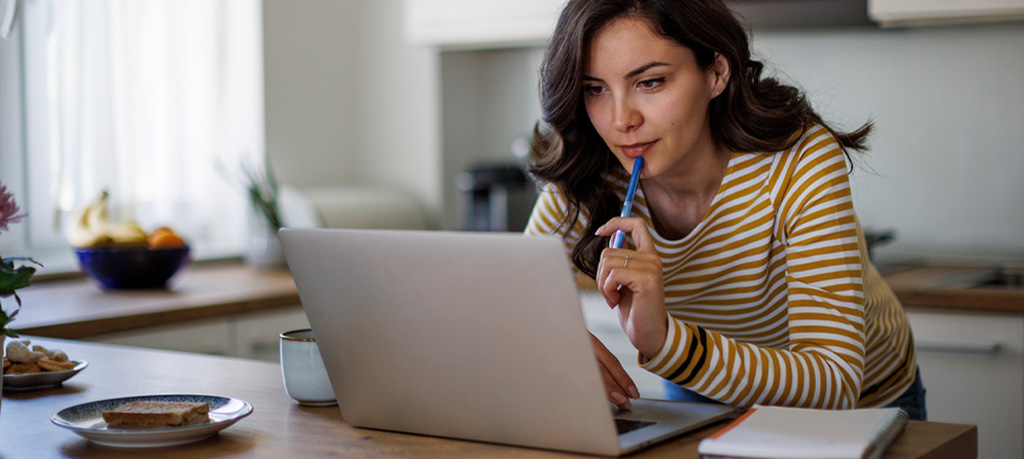 Woman in striped shirt using laptop computer to compare savings rates while sitting at kitchen table with coffee.