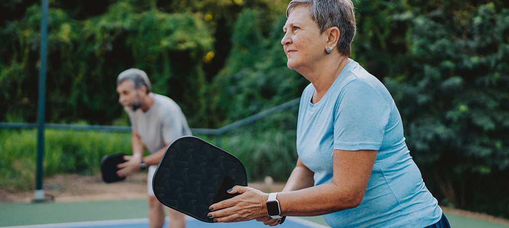 Two people play pickleball on an outdoor court, with one person holding a paddle and preparing to return the ball.