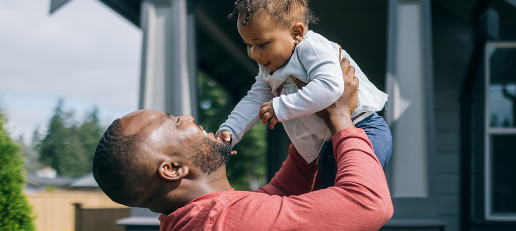 Joyful moment between father and child as dad lifts his smiling baby high overhead in front of their house. The father wears a red long-sleeve shirt and looks up at the baby with delight. The baby, dressed in a light blue outfit with hair styled in small buns, smiles down at their father against a backdrop of the home's dark exterior.