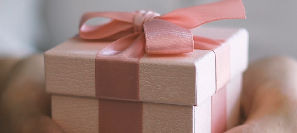 Close-up of hands gently holding a gift box wrapped in textured beige paper and tied with an elegant coral pink satin ribbon bow.