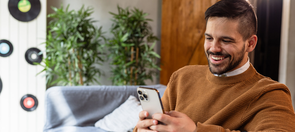 Close-up of a smiling man wearing a brown sweater, looking at his phone. He is seated on a gray couch with a pillow, and there's a plant in the background.