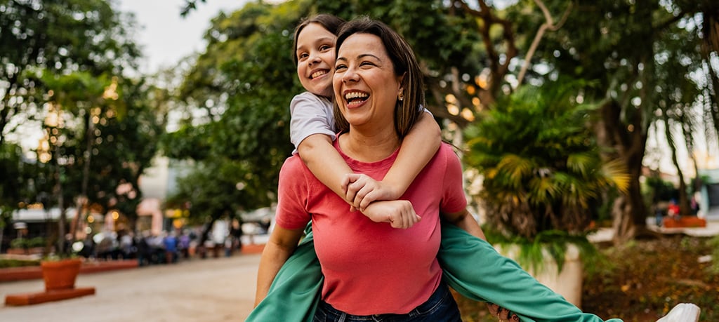 An adult carries a child on their back while walking along a path in a green park.