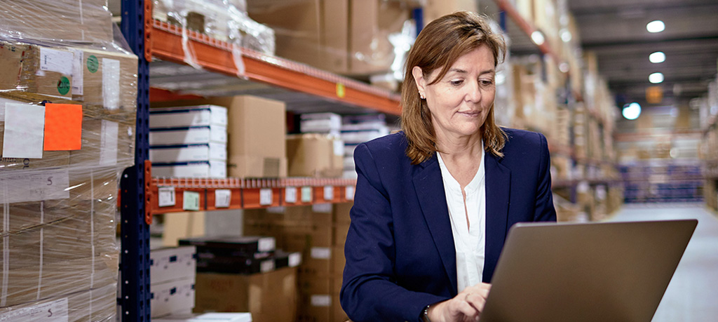 Professional woman in navy blazer using laptop in warehouse facility with orange shelving and inventory boxes in background