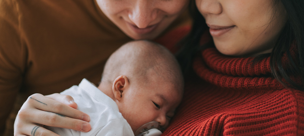 A couple leans close together, eyes closed, tenderly cradling their sleeping newborn baby wrapped in a white swaddle.