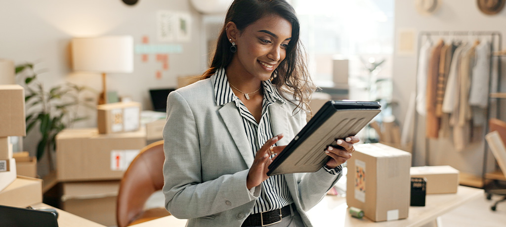 Smiling woman in a light gray blazer and black-and-white striped shirt holds a tablet in a bright office, with shipping boxes and clothing racks in the background.