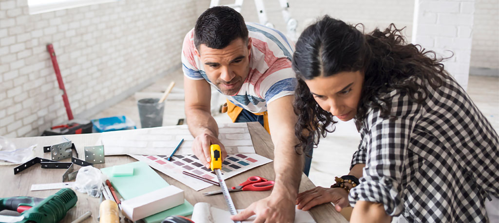 A man and woman work together on a home renovation, carefully measuring wallpaper and comparing color swatches. Tools and building materials are visible on the table around them.