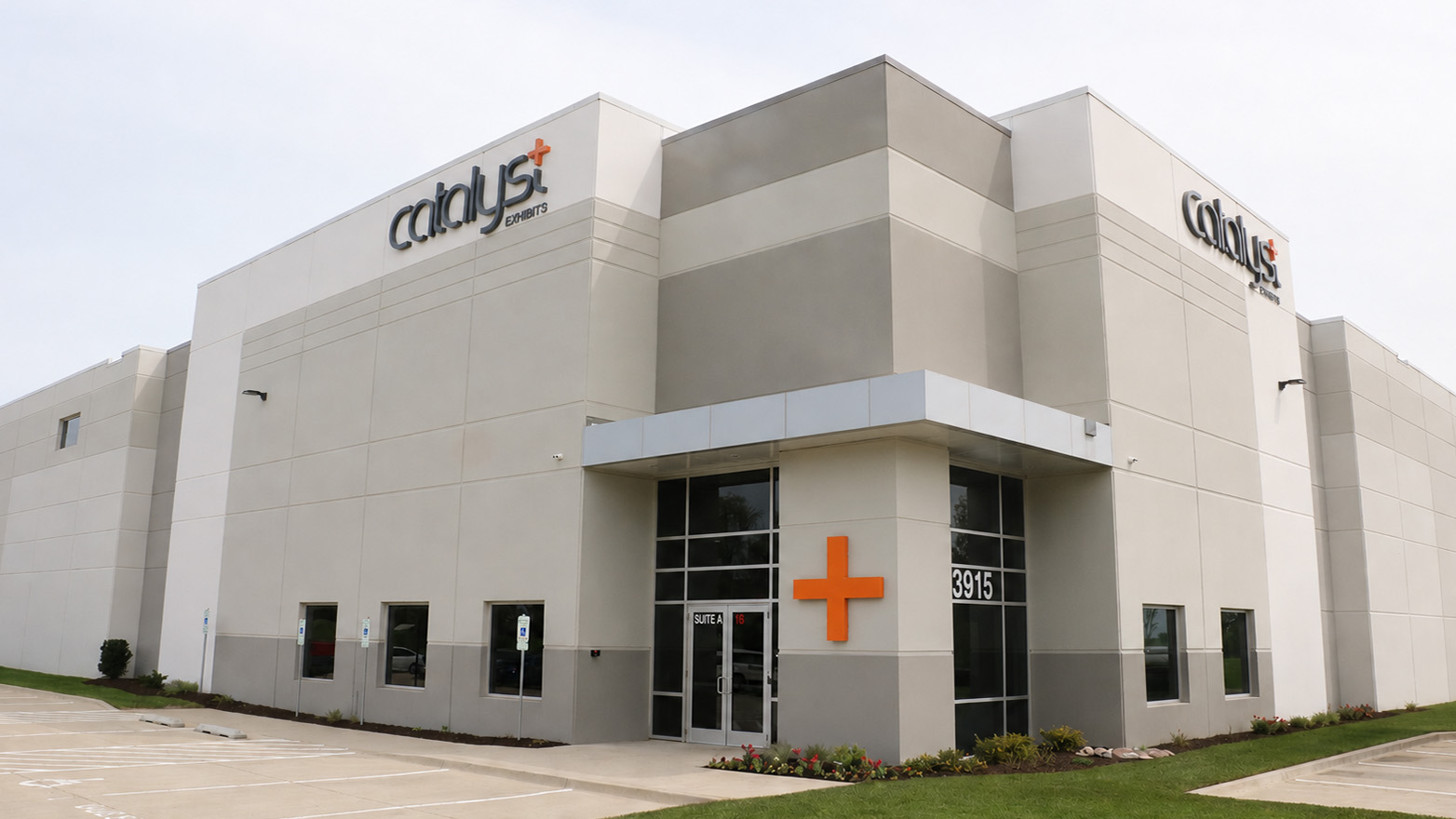 Exterior view of the Catalyst Exhibits office building featuring a modern beige and gray facade, large windows, the Catalyst logo on two corners of the building, and an orange plus sign displayed near the main entrance. A sidewalk, parking area, and landscaped lawn surround the building.