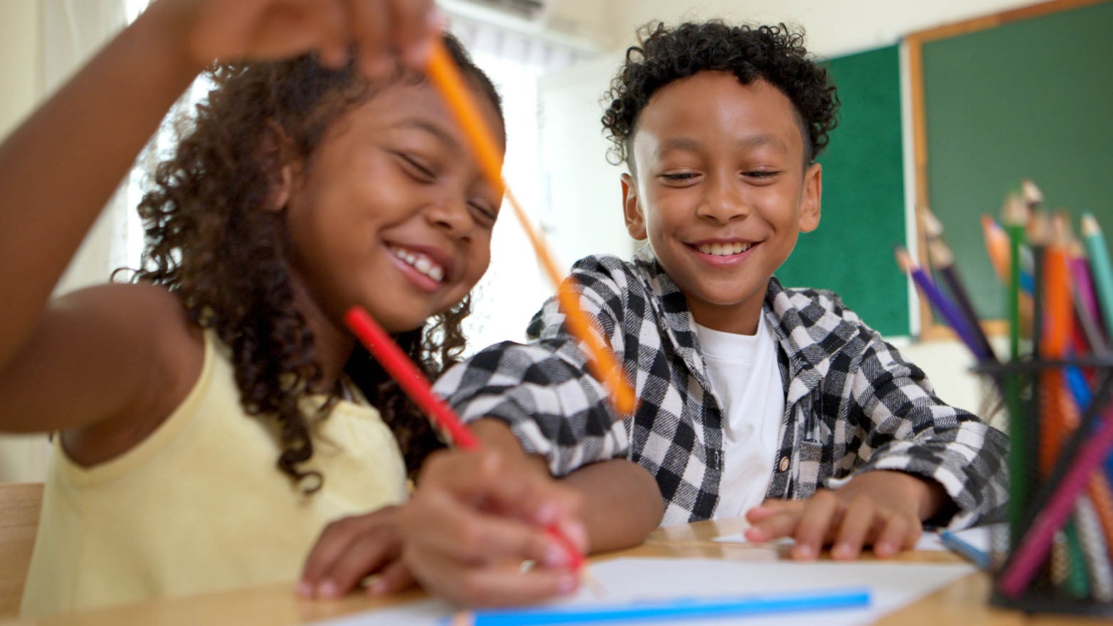 Two children sit at a classroom table drawing with colored pencils on paper. A container of colored pencils is visible in the foreground, with a chalkboard and classroom setting in the background.