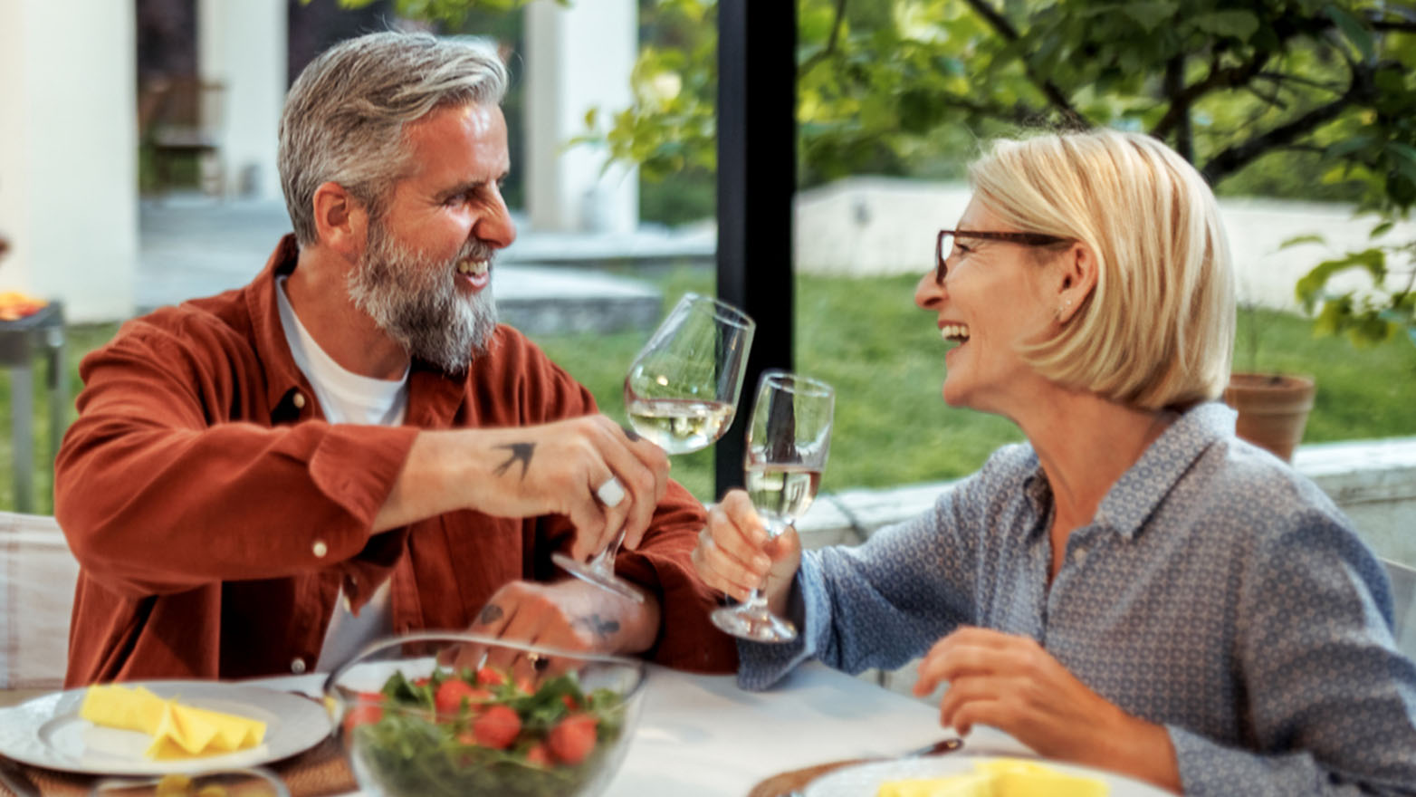 Two people sit at a dining table, raising glasses of white wine during a relaxed meal.