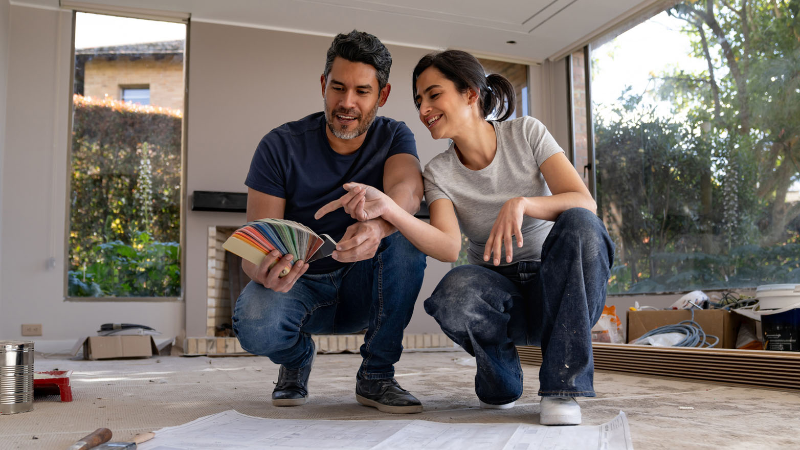 Happy couple crouching on an unfinished floor in their home, examining a fan of colorful paint swatches together during a renovation project, with large windows and construction materials visible in the background.