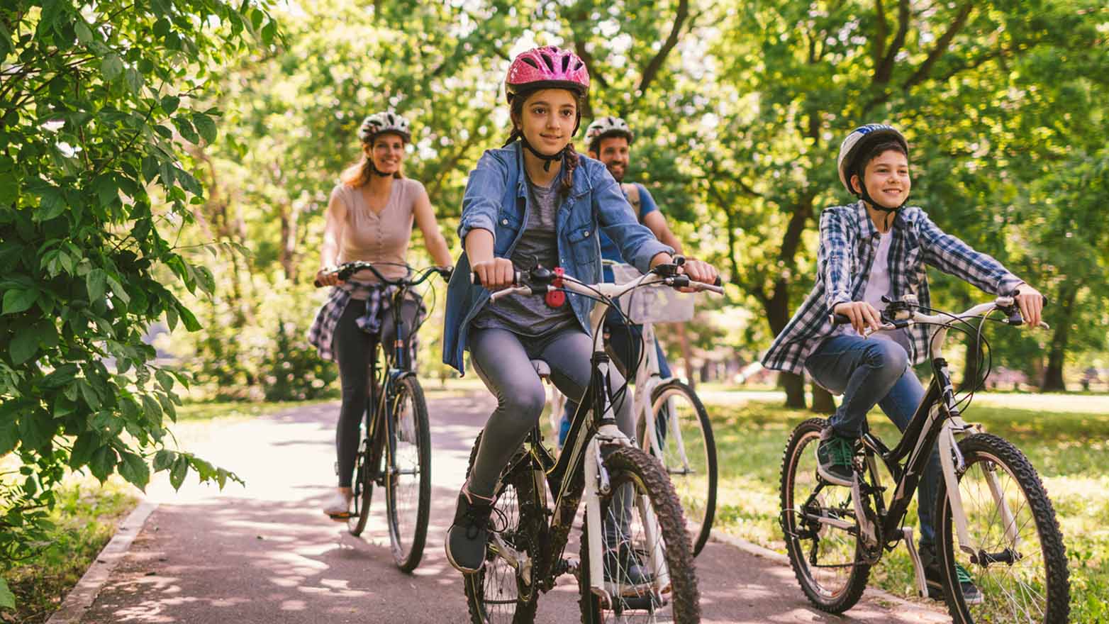 A family of four wearing helmets smiles while riding bicycles on a sunny, tree-lined park path.