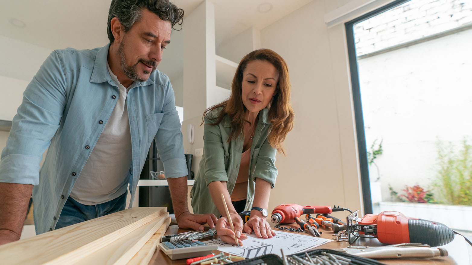 A man and woman lean over a table, planning a home project. The woman draws on paper, surrounded by wood planks and a power drill.