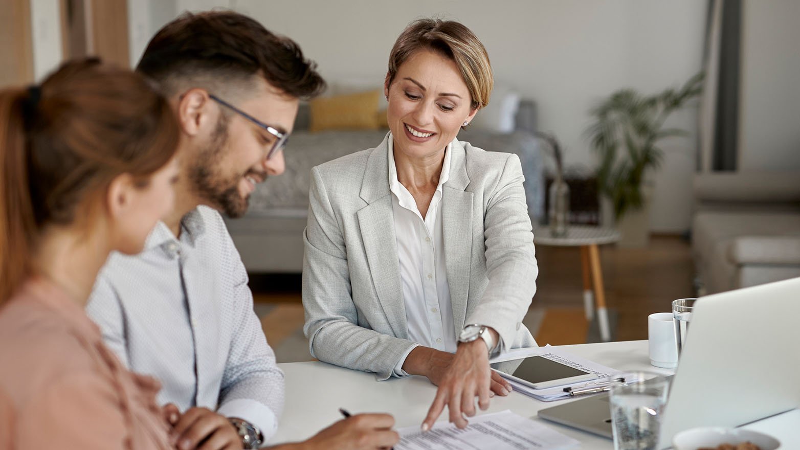 Female advisor in a gray blazer smiles, pointing to a document for a young couple reviewing it at a desk.