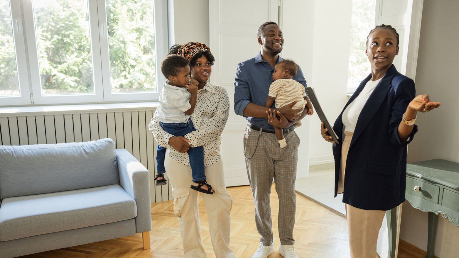 Real estate agent gestures in a bright, sunlit room to a smiling family of four.
