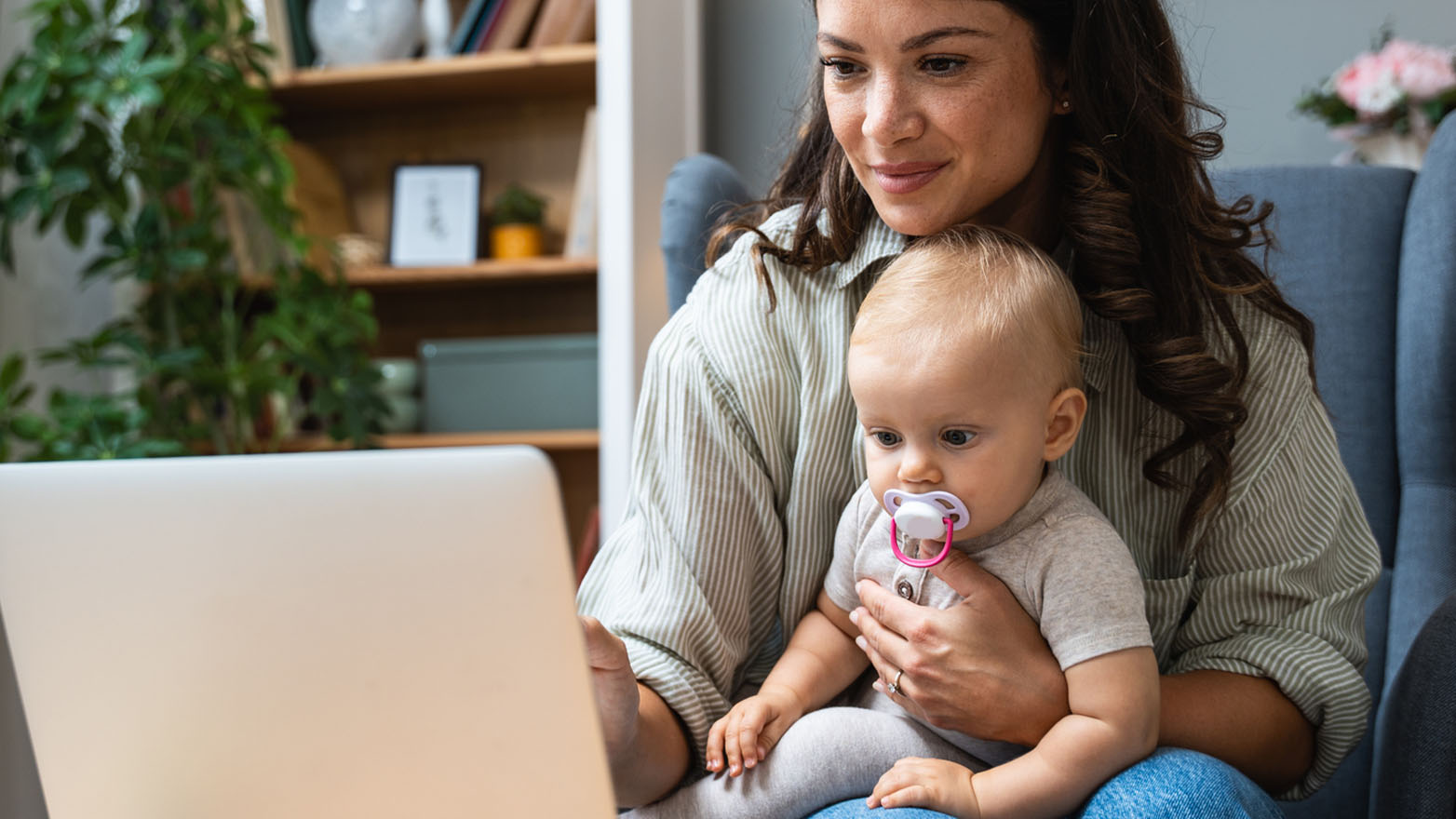 A mother sitting in a chair with her baby in her lap, both looking at a laptop screen. The woman is wearing a striped shirt, and the baby has a pacifier.