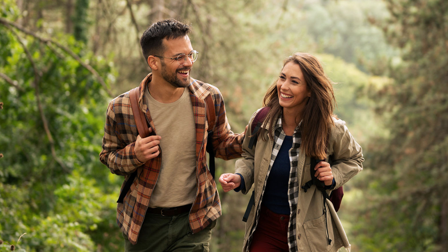 A young man and woman smile and laugh while hiking on a trail in a lush green forest.