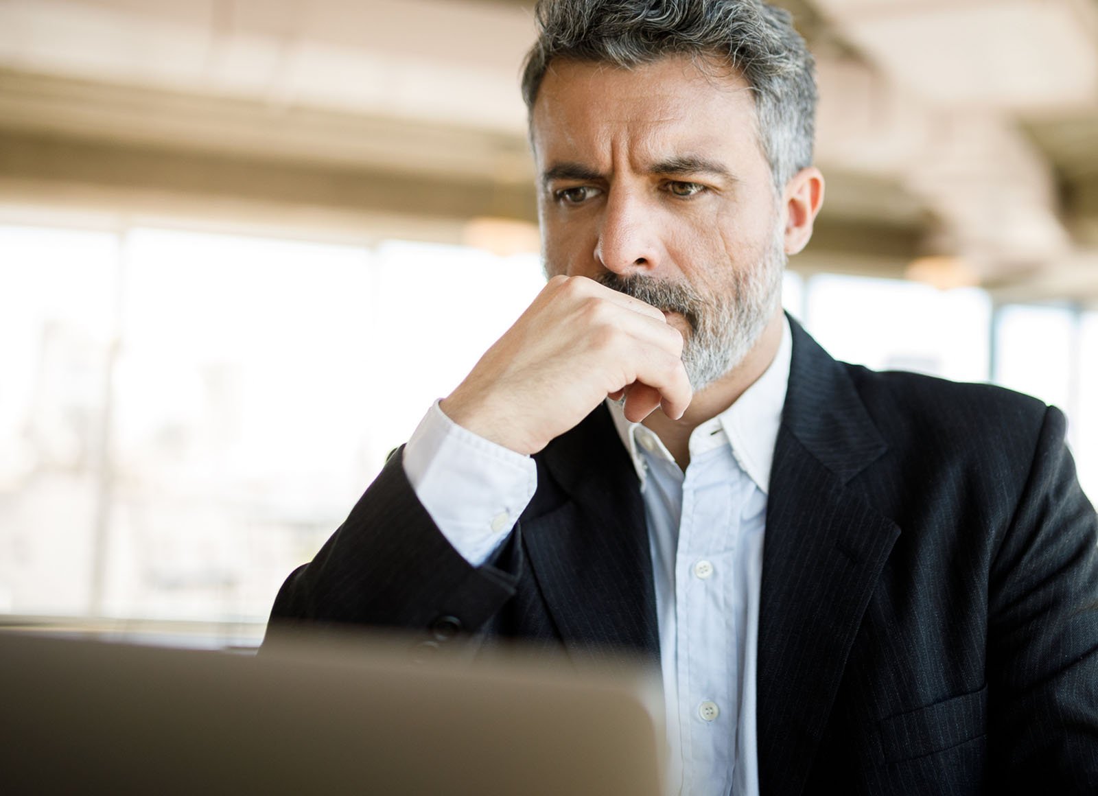A man in a business suit looks contemplative, his face cradled by his hand as he gazes at the screen of his laptop.