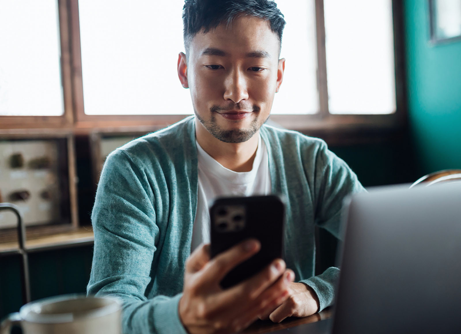 A person sits at a café table, holding a smartphone while working on a laptop.