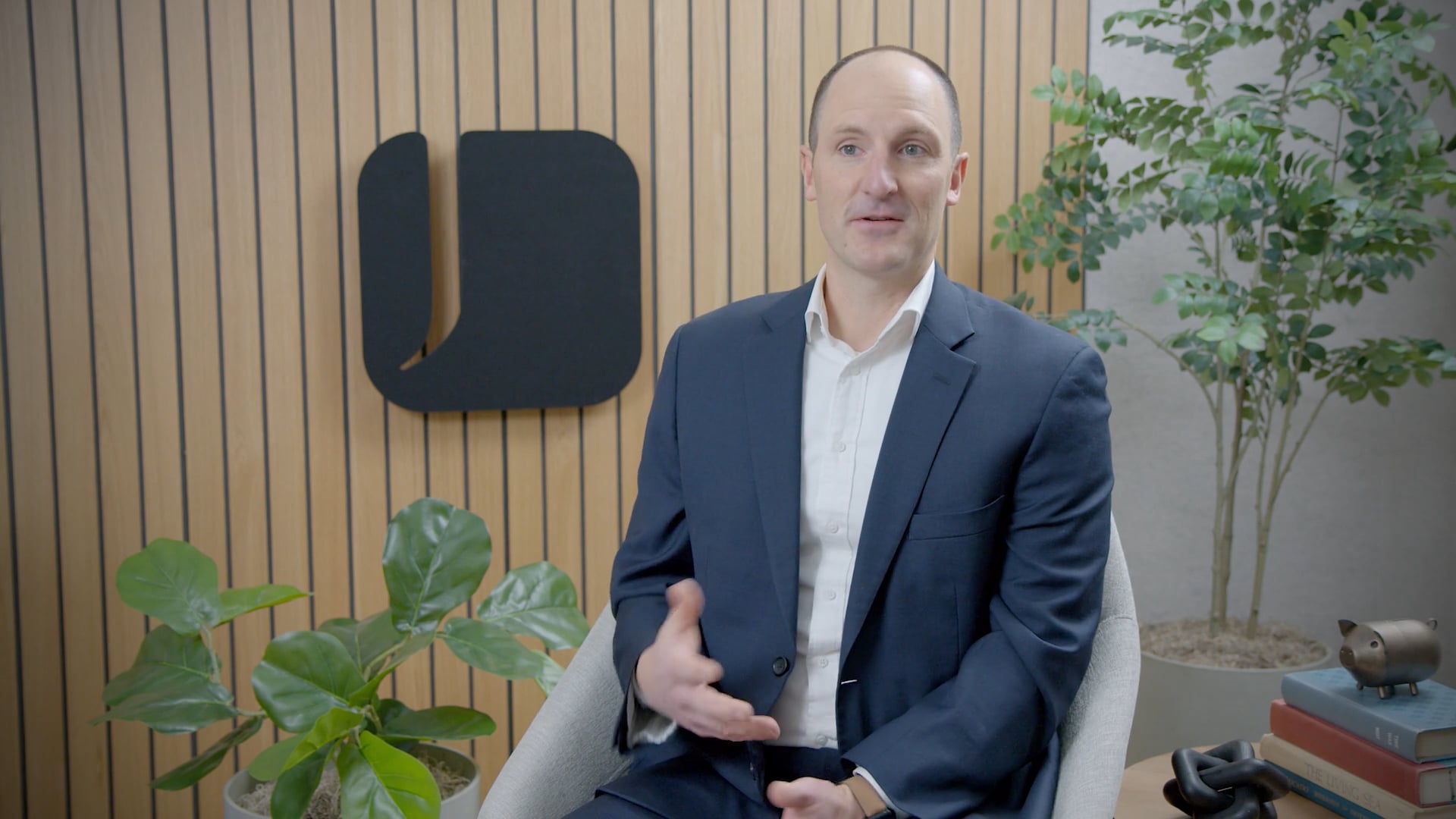 A man in a blue suit and white shirt sits in a gray armchair, speaking and gesturing in a modern office with a wood-paneled wall behind him.
