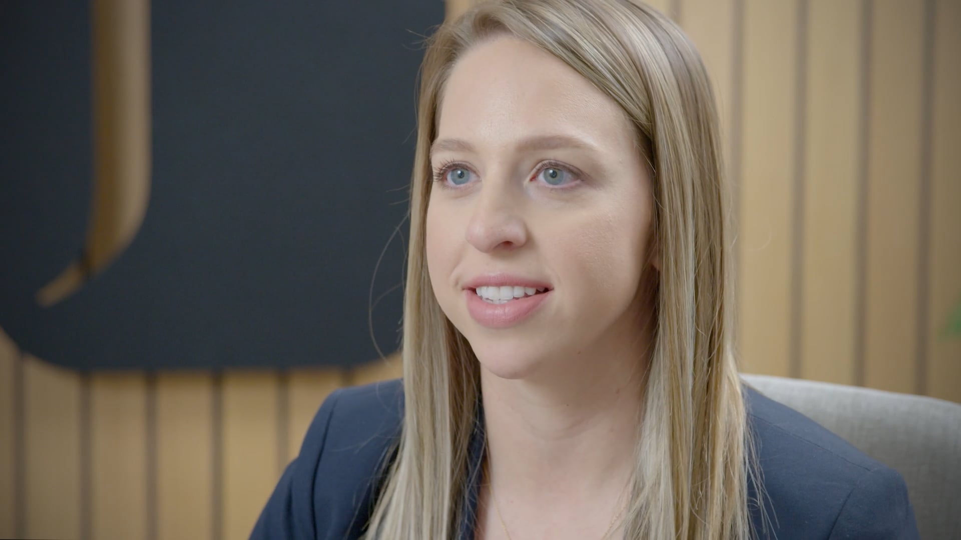 Close-up of a young woman with blonde hair, blue eyes, and a gentle smile, wearing a dark blazer.