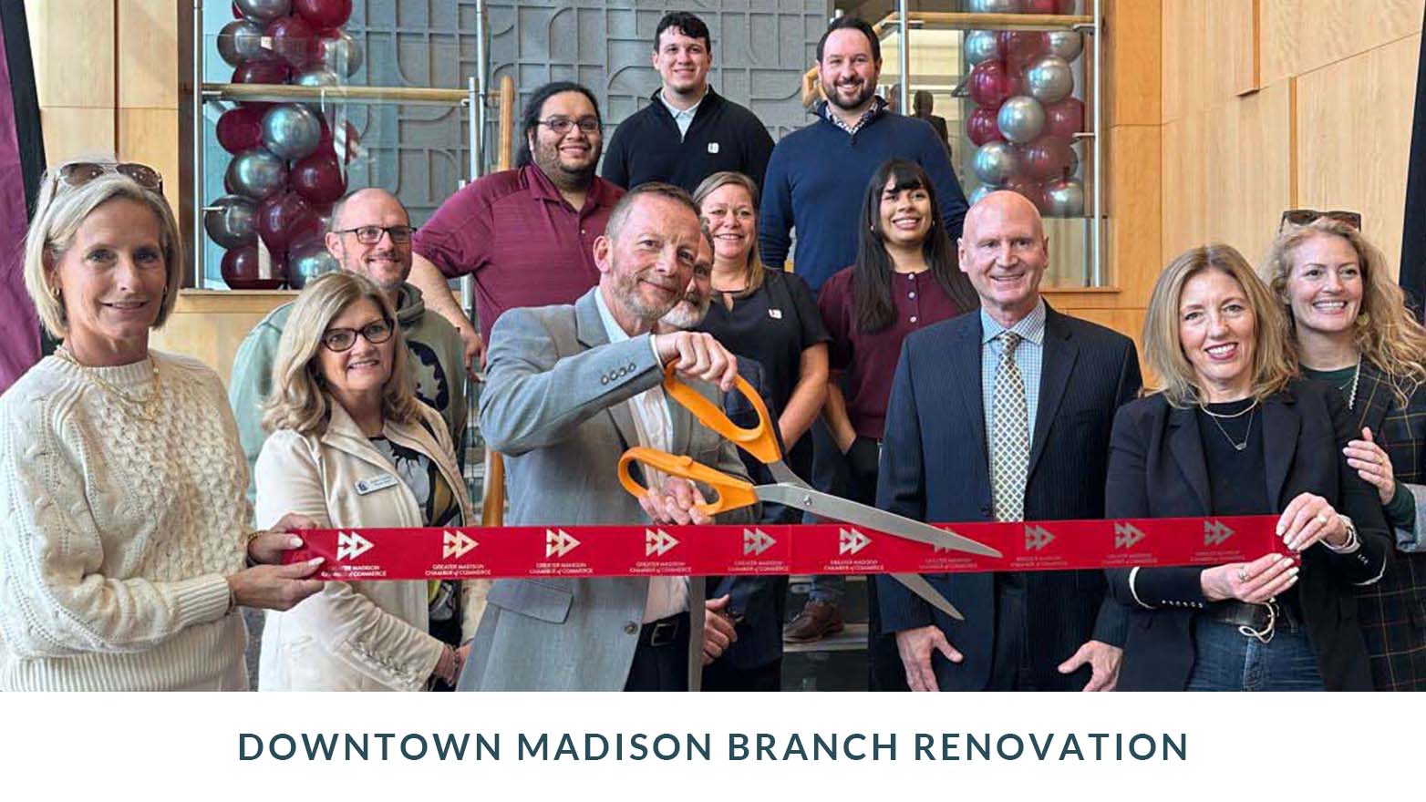 Group photo taken indoors during a branch renovation event, showing a group of people standing together while one person cuts a red ribbon with oversized ceremonial scissors. Caption below reads “Downtown Madison Branch Renovation.”