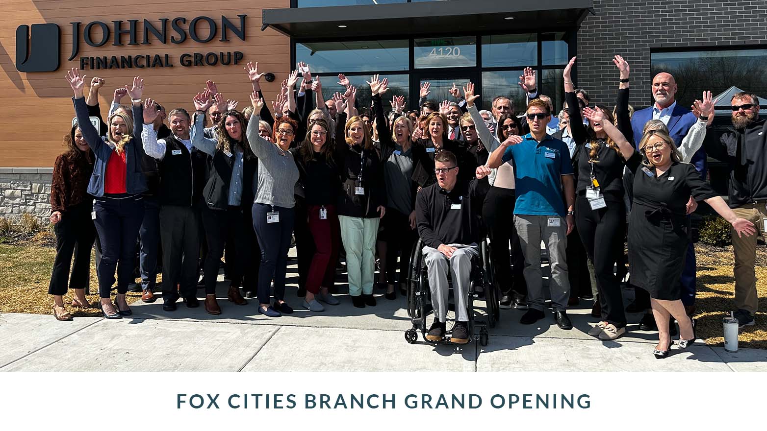 Group photo taken outdoors in front of a Johnson Financial Group building during a branch grand opening, showing a large group of people standing together with arms raised. Text below reads “Fox Cities Branch Grand Opening.”