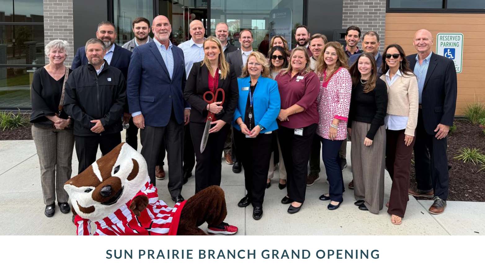 Group photo taken outdoors in front of a building entrance during a branch grand opening, showing a large group of people standing together; one person holds oversized ceremonial scissors, and a mascot in the foreground wears a striped shirt.