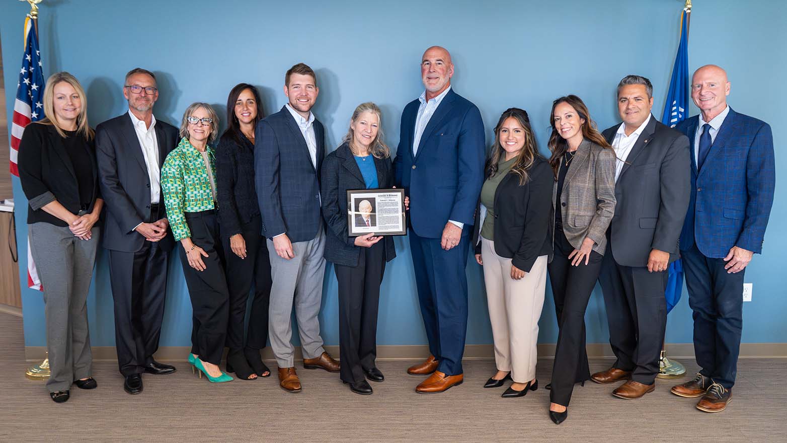Group of twelve people standing in a line indoors against a blue wall, with U.S. and Wisconsin flags visible at either side; one person near the center is holding a framed plaque.