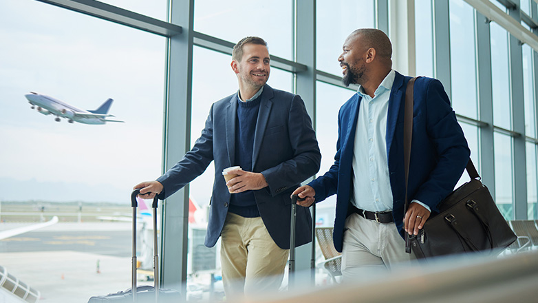Two businessmen in suits, carrying luggage, walk and talk in an airport terminal as a plane takes off in the background, visible through a large window.