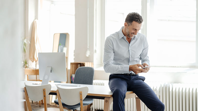 Man standing next to his desk looking at his mobile phone.