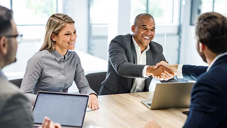 Four people sit at a table, two men are shaking hands while two other people, a woman and a man, look on. There are two laptops on the table, along with a pen and notepad.