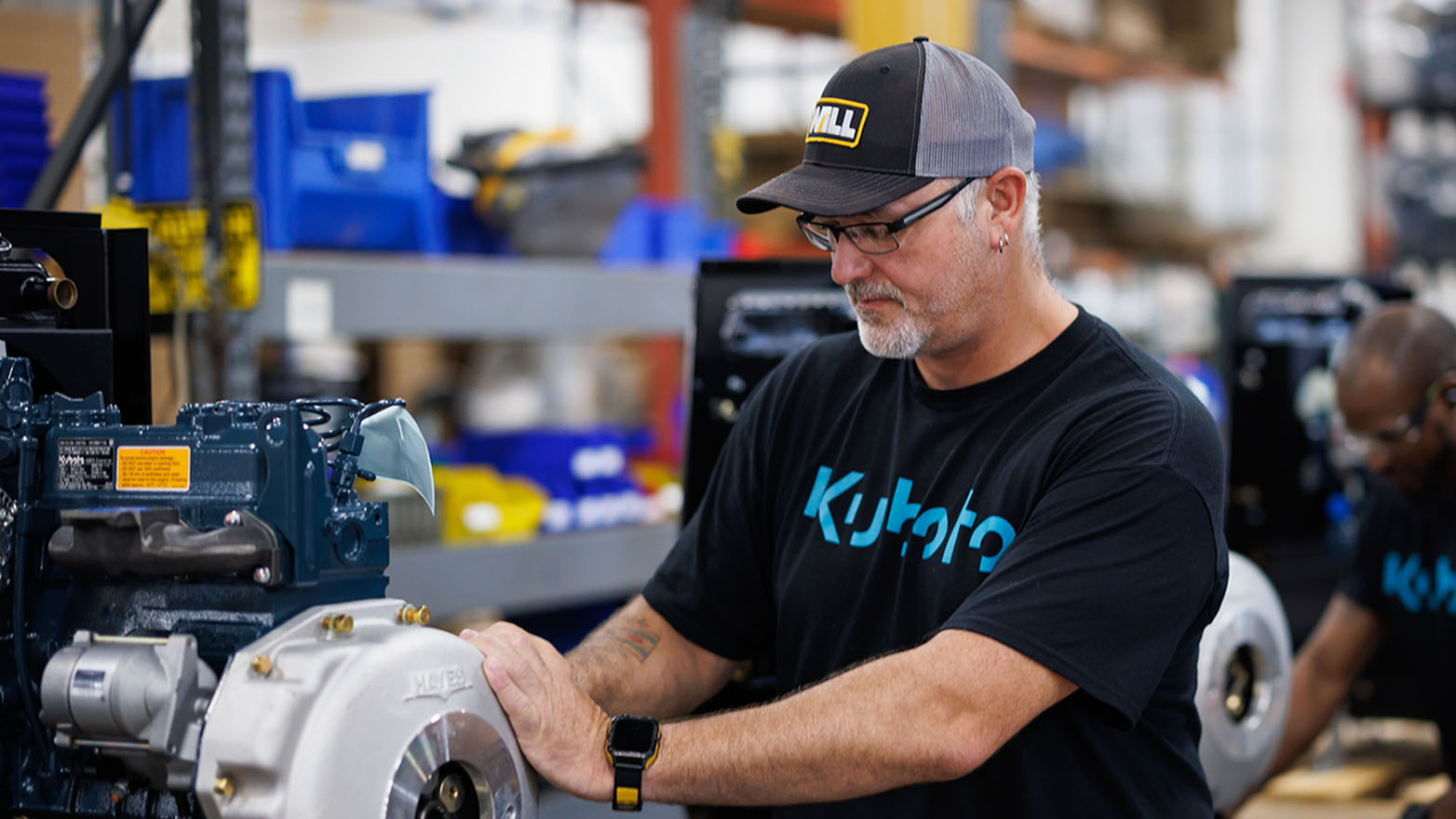 A man wearing a Kubota shirt is crouched over, performing maintenance on a engine.
