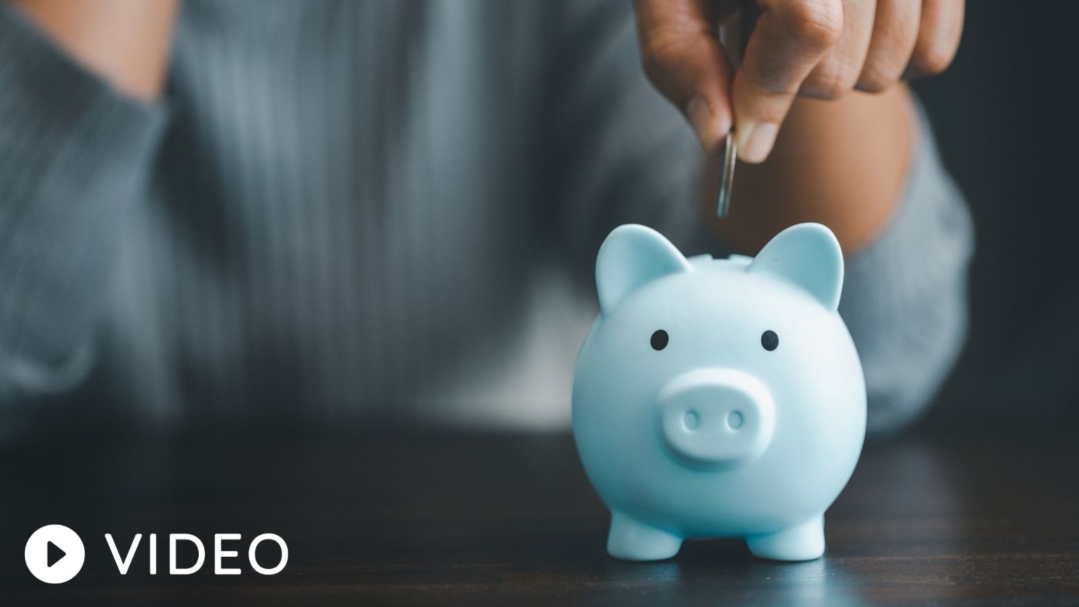Close-up of a hand dropping a coin into a light blue piggy bank on a dark wooden table.