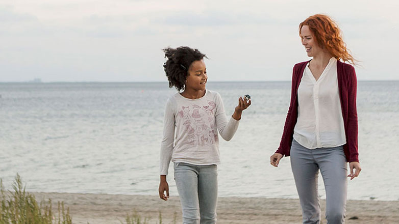 Mom and daughter walking barefoot together on a sandy beach.