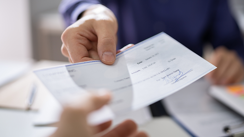 A person's hand is handing a check to another person, the other person's hand is blurry. The check is filled out and signed.