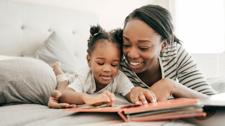 Mom reads her toddler daughter a book while they lay on the bed.