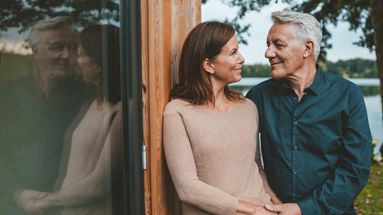 A mature man and woman, embracing gently, gaze into each other's eyes. They are standing near a wooden structure by a lake.