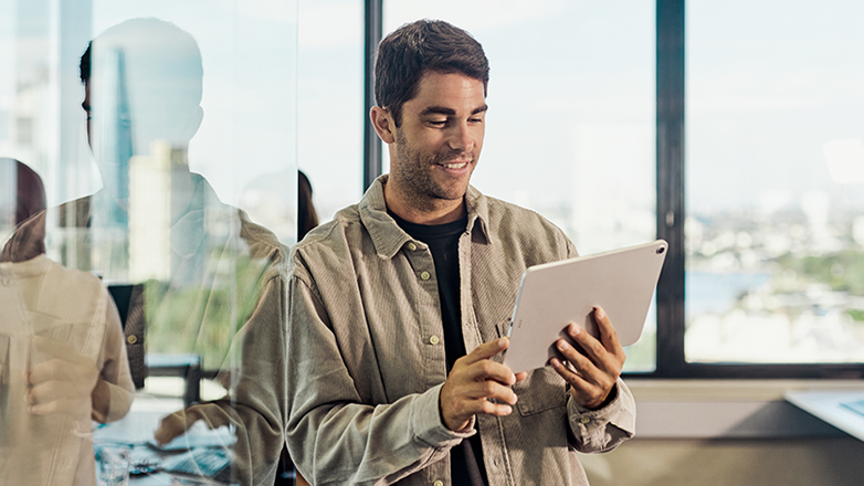 A man stands in an office, looking at a tablet in his hands. He is smiling, and the office has large windows looking out to the city. The image suggests a positive and professional work environment.