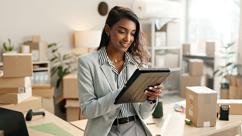 A young woman stands in an office with boxes stacked behind her. She looks down at a tablet, checking details and smiling. The background is out of focus, showing a cluttered office space with more boxes, a potted plant, and a desk with a computer. The woman's smile suggests she is confident and content with her work.