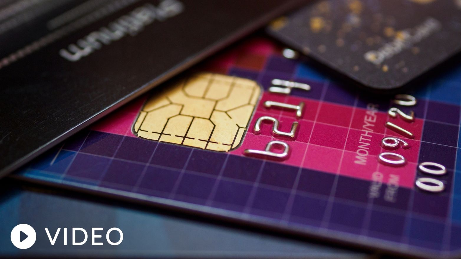 Close-up of several stacked credit cards, with a purple and pink card in front showing a gold chip and raised silver numbers. A black platinum card is partially visible on top.