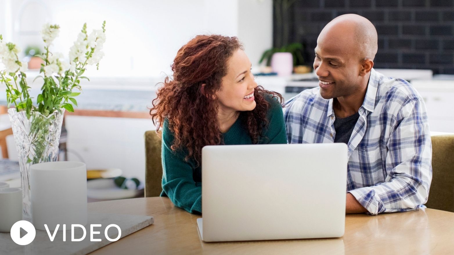 A man and woman sit at a wooden table, smiling at each other with an open laptop in front of them.