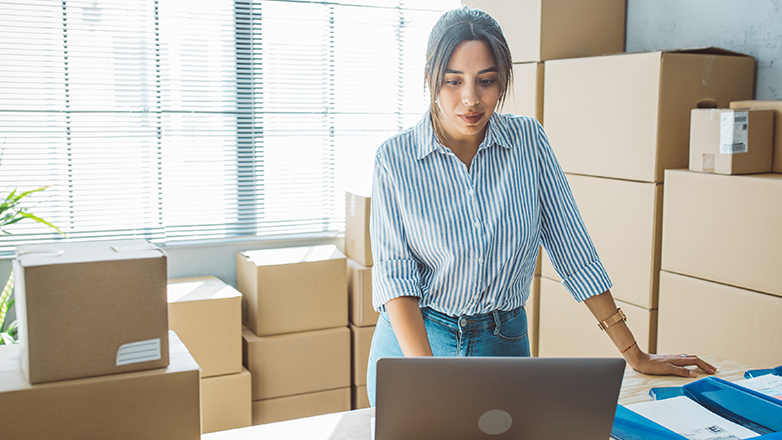 A young woman stands in a room full of boxes. She is wearing a blue and white striped shirt and jeans. She is looking at a laptop on a table.