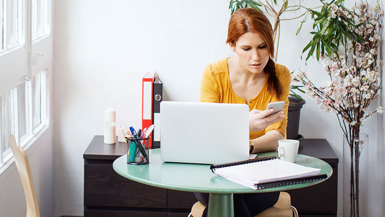 Woman sitting at her office table working on a laptop and checking her mobile phone.