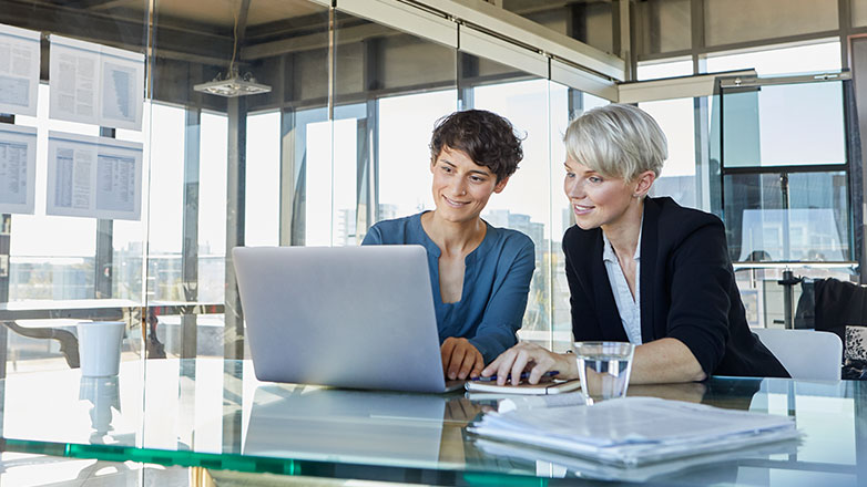 Two female executives sitting at a desk reviewing laptop.