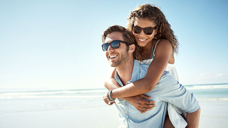 Husband gives wife a piggyback ride at the beach on a sunny day.