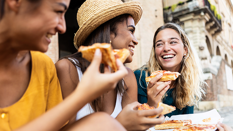 Three young women are sitting on the ground and eating pizza. They are all smiling and laughing.