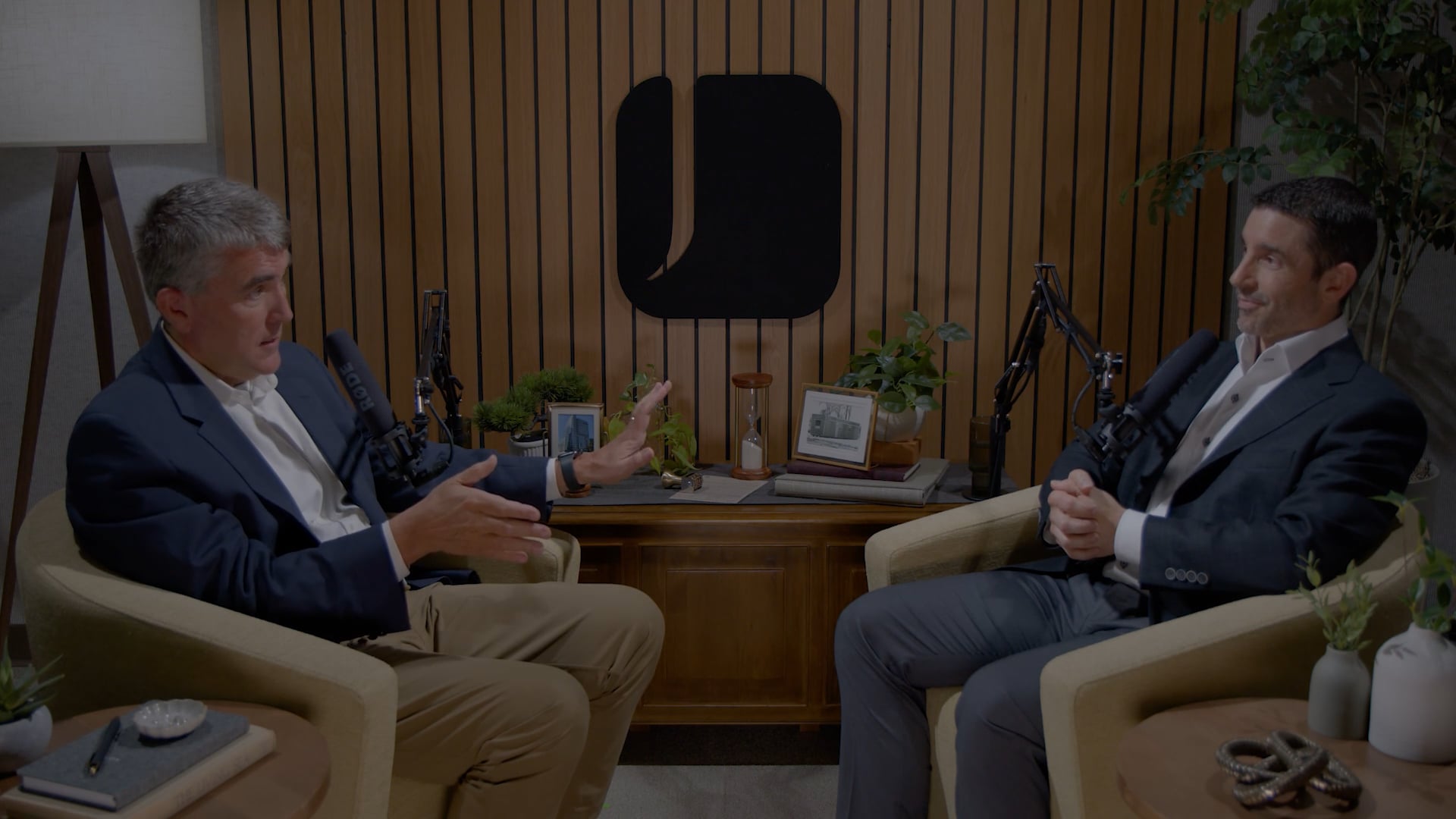 Two people seated in armchairs recording a podcast interview, speaking into microphones in a studio with wood-paneled walls, plants, and a table between them.